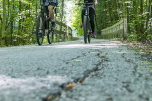 Zwei Radfahrer:innen im Wald und Risse im Asphalt